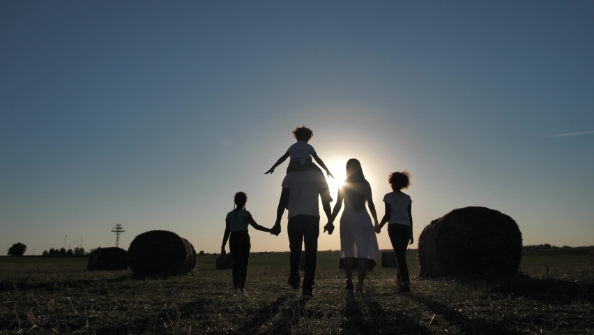 Silhoutte of multinational family, african american man, caucasian wife and two mixed race preteen daughters enjoying walk hand in hand on wheat field, youngest girl riding on dad's shoulders