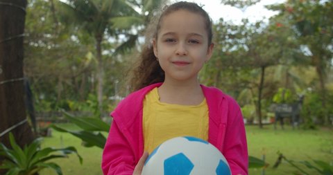 A cute little girl throws a soccer ball upstairs and catches it with her hands, smiles and looks at the camera, prepares to play with the ball, stands against the backdrop of palms, trees and sunlight