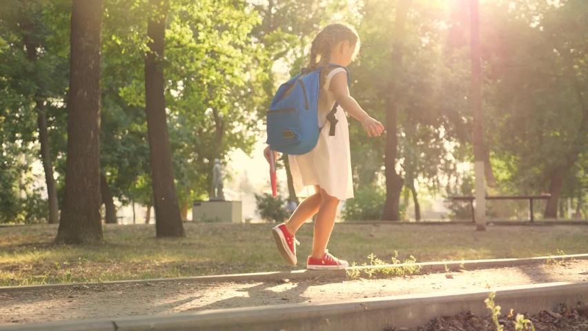A little schoolgirl with a backpack and a book goes home through the Park. Rear view. A child hurries to the Playground after school. A happy kid plays in the Park after school. Royalty-Free Stock Footage #1066983859