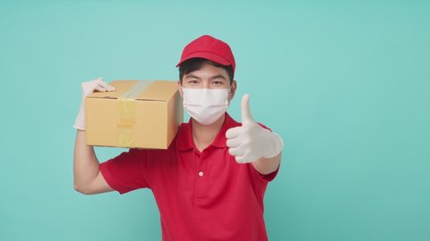 Isolate background, Asian delivery man wearing mask in red uniform holding medium box parcel in his shoulder to deliver to customer. The male postman show thumbs up and cheerful face and smile.