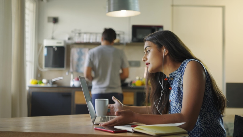 An angry and frustrated Indian female corporate entrepreneur is shouting at office colleagues during an online video conference meeting while her husband is preparing a cup of coffee in the kitchen Royalty-Free Stock Footage #1066996930