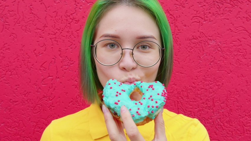 A young woman eating a donut. A female hipster with green hair against a red wall. Contrasting saturated colors | Shutterstock HD Video #1066998085