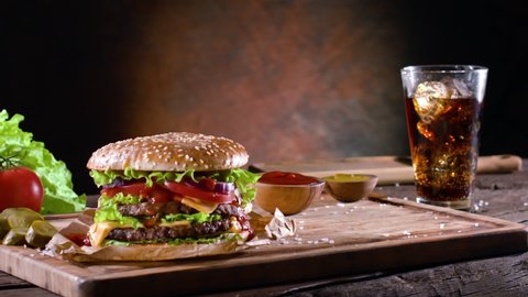 French fries and salt slowly land on top of the wooden board with hamburger and cola. Behind a dark brown background. 