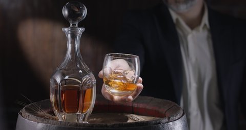 Man in a suit and beard (sommelier) takes a glass of whiskey on ice from a vintage whiskey barrel and shakes the glass to spread the smell of whiskey. Slow motion 4K, 150 fps, Blackmagic Ursa Pro G2. 