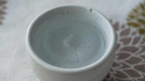 A small cup of pottery containing Japanese sake. Drop fall and bounce to create ripple. Japanese pattern tablecloth of chrysanthemum flower in the background. Asian alcoholic beverage made from rice