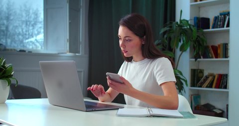 Young female student prepares homework using mobile phone laptop indoors slow motion. Casual woman searching information for project in smartphone. Freelance home education coronavirus quarantine 