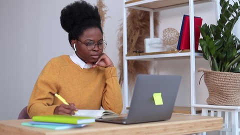 African American Millennial woman in yellow sweater, wearing headphones, glasses, sitting at her computer watching a webinar, taking notes in her notebook. Woman taking online course, studying, workin