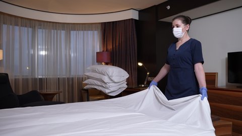 Diligent female hotel worker carefully spreading clean white sheet by tossing while making bed for guests. Neat maid in uniform, medical mask and gloves preparing bed in hotel room during coronavirus