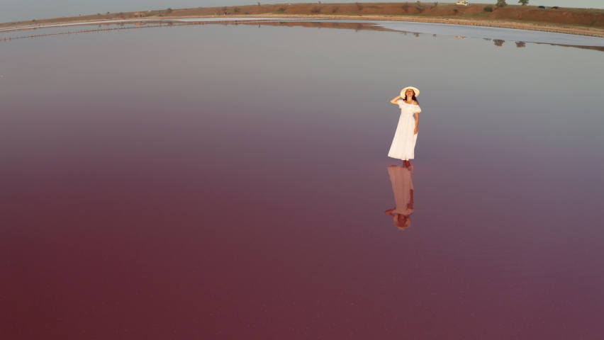 Beautiful woman in a white dress walking on a pink lake at sunset