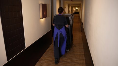 Back view of female hotel room cleaner walking with cleaning trolley along long corridor stretching into distance. Chambermaid with janitor cart going to clean hotel room before guest's arrival