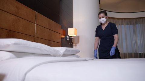 Neat chambermaid in mask, gloves and uniform carefully straightening blanket after making bed with crisp white linens in hotel bedroom. Diligent maid preparing room for hotel visitors during covid-19