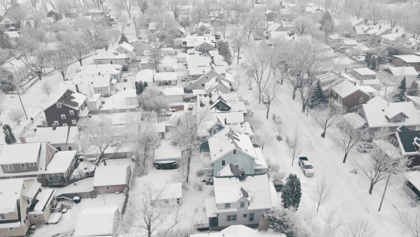 Aerial video flying over a white snow swept neighborhood featuring snowy roofs, yards, and streets.