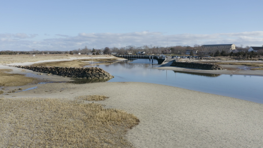 The camera pushes in and over the breakwater structure. This is the entrance to Rock Harbor in Orleans, MA. People can be seen near the boat ramp and public beach. The low tide reveals the boat ramp.