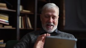 Close-up view of happy smiling mature adult male greeting and waving hand, talking by digital tablet. Aged man alone at home has video call on tablet to keep in touch with family during isolation.
