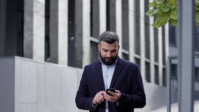 Handsome businessman using smartphone standing near business center. Happy smiling young man using mobile phone apps, texting message, browsing internet, watching video,tapping,looking at smartphone.