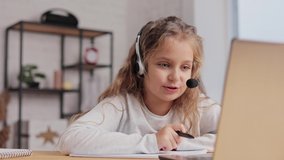 Schoolgirl in headset has online lesson, video call with teacher, answering questions,check of knowledge.Cute primary school girl studying at home using laptop. Distance learning. Home education.