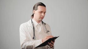 A male doctor with long hair in a medical gown writes on paper on a clipboard. Gray background.