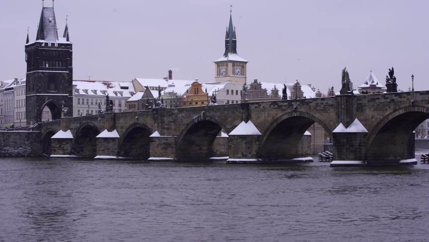 view of the snow-covered Charles Bridge on the flowing river Vltava and snow-covered roofs of buildings in the old town of Prague during the day in winter and there are silhouettes of pedestrians Royalty-Free Stock Footage #1067045176
