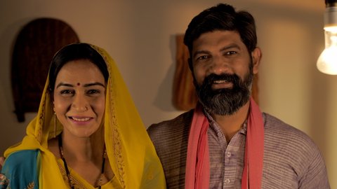 Affectionate Indian village couple standing indoors while smiling for the camera. Closeup shot of a traditional woman in Saree and man in Kurta-Pyjama during the night - Happy nuclear family