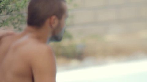 Sexy confident guy sitting near the pool, someone puts hand on his back , slow motion