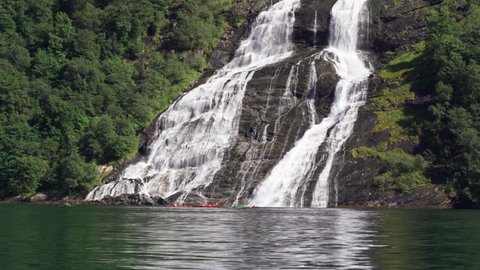 One of the streams of the Seven Sisters waterfall in Geiranger fjord. Water cascading over the dark rocks joining with the fjord. The group of kayakers passing by admiring the view.