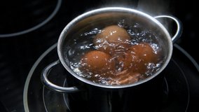 HD video. Selective focus. Close-up view of four brown chicken eggs boiling in hot water in small stainless saucepan on black kitchen stove (induction cooktop). Easter preparation theme.