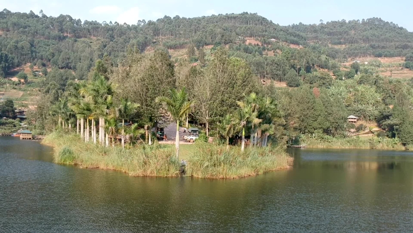 Camera pushing in from the water of a lake to a small peninsula showing a palm tree in the center and trees all around. The water is calm and there are green hills all around. Light blue skies above.