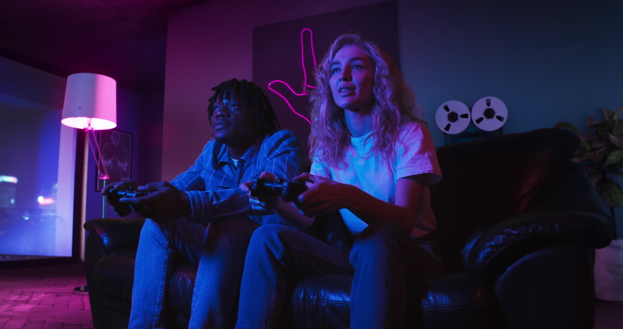 Young man and woman are playing video games together using controllers. They are sitting on the sofa in the living room with red and blue neon lighting. Girl wins and celebrates victory. Royalty-Free Stock Footage #1067074444