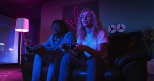 Young man and woman are playing video games together using controllers. They are sitting on the sofa in the living room with red and blue neon lighting. Girl wins and celebrates victory.