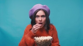 stressed woman in beret holding bowl and eating popcorn on blue
