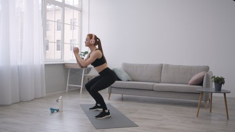 Home Workout. Athletic Woman Doing Jump Squats Exercising Wearing Headphones Indoor. Side View. Legs Exercises, Female Training Concept. Side View, Slow Motion