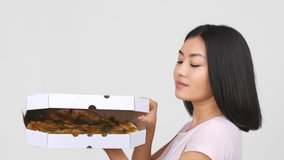 Asian Woman Opening Pizza Box And Smelling Delicious Italian Street Food, Holding Paperboard Box Near Face And Smiling To Camera Posing Over White Background. Tasty Junk Food. Slow Motion
