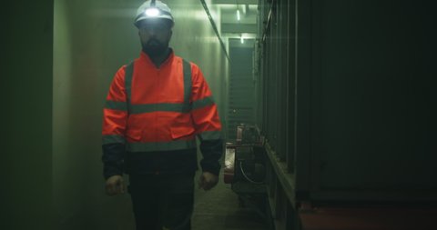 Pan left view of man in uniform and hardhat walking in dim hallway and examining machine during work on factory