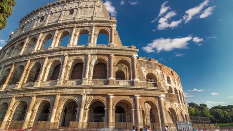Many tourists visiting The Colosseum or Coliseum timelapse hyperlapse, also known as the Flavian Amphitheatre in Rome, Italy. Orange light at sunset. Blue cloudy sky at sunny day