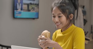 Young beautiful asian woman eat hamburger while work or study online with laptop computer. Happy girl smile in yellow casual clothes at house during quarantine. Social distance new normal concept.