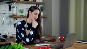 Beautiful woman chef teacher in beige apron teaches online remote video call conference laptop computer, listens housewife tells students shows ingredients for making salad to webcam in home kitchen