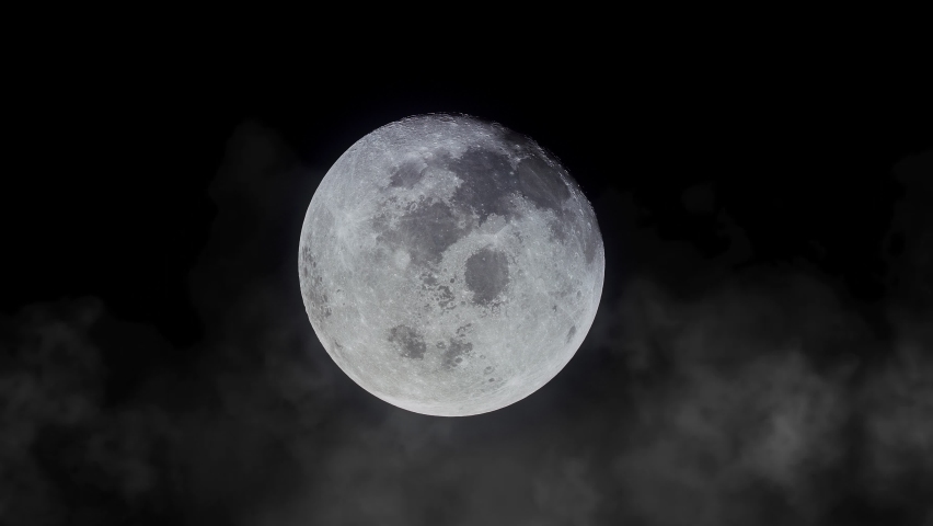 The bright full moon on the dark sky with clouds being blown by strong winds during a storm at night. Close-up of the glowing moon in the darkness during windy and cloudy weather. Midnight 4k video.