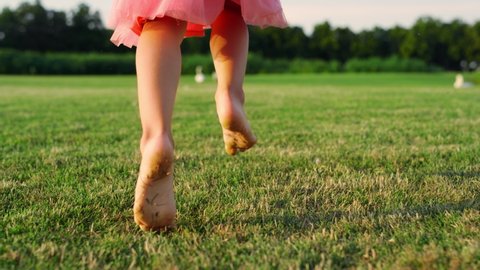 Unrecognizable girl walking barefoot in city park. Unknown girl in pink dress playing on green grass. Rear view of female kid legs running at sunrise meadow.