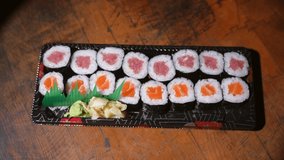 This video shows a close up, top view of anonymous hands using chopsticks to take pieces of delicious salmon and tuna sushi rolls from a carry out dish on a wooden table background.