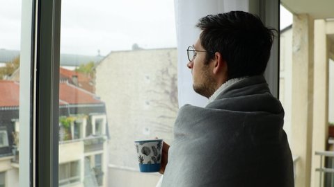 Young man resting at home and looking through window during overcast day with cosy blanket and cup