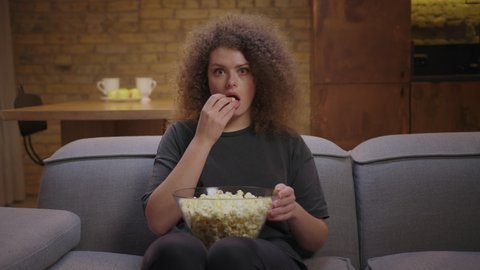 Open-eyed young woman watching TV and eating pop corn sitting on couch at home. Female shows wow emotion while watching movie or tv show.