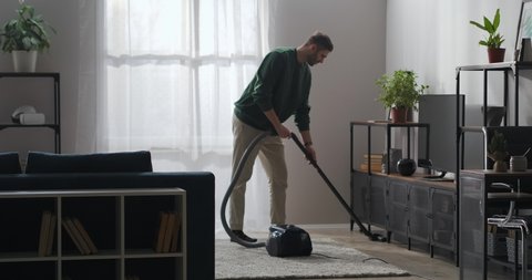 married man is doing clean-up at home, using vacuum cleaner for cleaning carpet and floor in living room