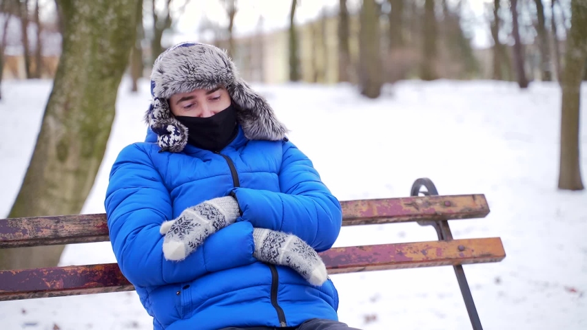 Portrait of a tired young man sitting on a bench closed his eyes in cold winter weather. A young adult man sleeps in the park in a mitten hat and a protective medical face mask. Exhausted resting