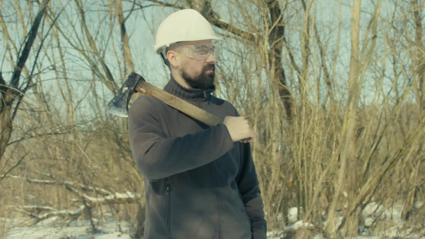 Portrait of a Caucasian bearded lumberjackwitg ax. Smiling woodcutter in a hardhat and glasses. Working in the forest in winter. Rustic. 