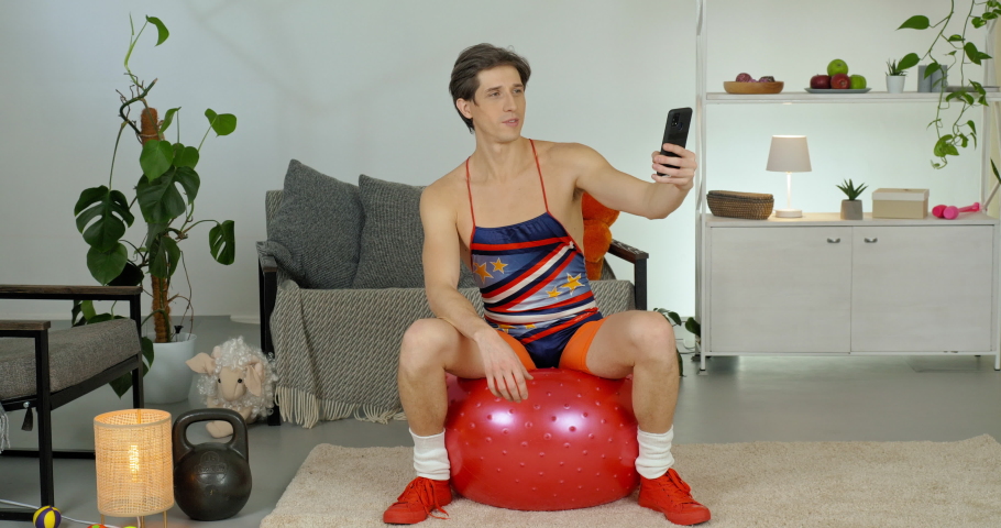 Funny caucasian man athlete sexy man in retro suit sitting jumping on fit ball holds smartphone in hand comically posing puts thumb up shows two fingers peace symbol makes self photo on phone camera