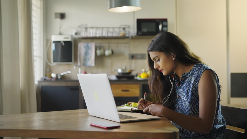 A young beautiful woman is sitting on a chair and writing down or taking down notes while attending an online video class or office meeting on a laptop in an interior house. Work from home concept Royalty-Free Stock Footage #1067145055