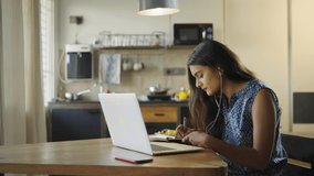 A young beautiful woman is sitting on a chair and writing down or taking down notes while attending an online video class or office meeting on a laptop in an interior house. Work from home concept