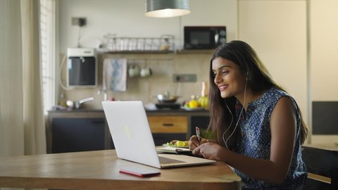 A young beautiful woman is sitting on a chair and writing down or taking down notes while attending an online video class or office meeting on a laptop in an interior house. Work from home concept