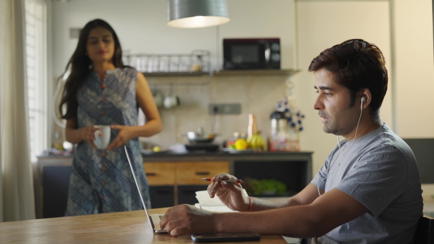 A shot of a husband is busy talking in an online conference meeting on a laptop while the supportive wife is offering him a cup of coffee in an interior house setup. Healthy relationship concept Royalty-Free Stock Footage #1067145058