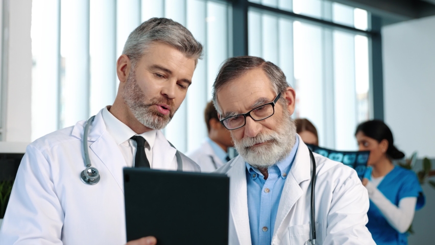Healthcare and modern medicine. Serious senior doctor looking together with his colleague at the survey results showing at the digital tablet while preparing for serious treatment at the clinic, a Technology Video by M_Agency